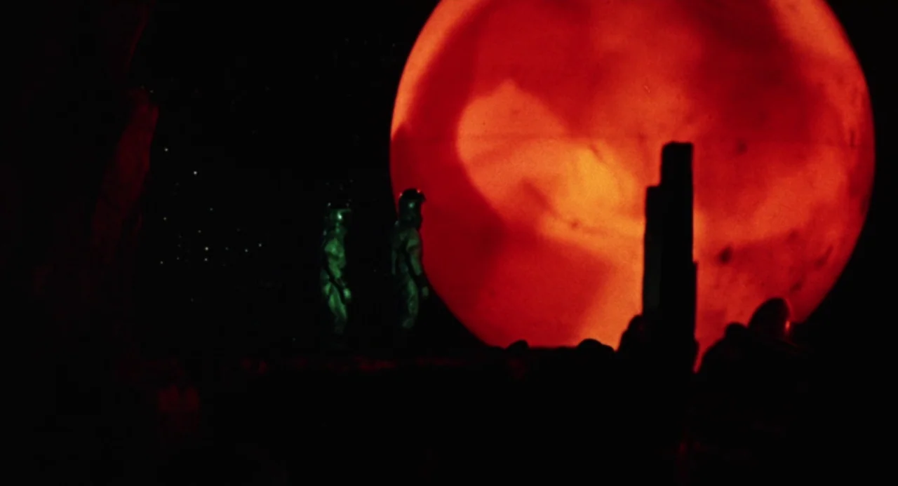Queen of Blood (1966) movie still: wide — A dark, craggy landscape features rock formations under a black starry sky. Two figures in suits stand…
