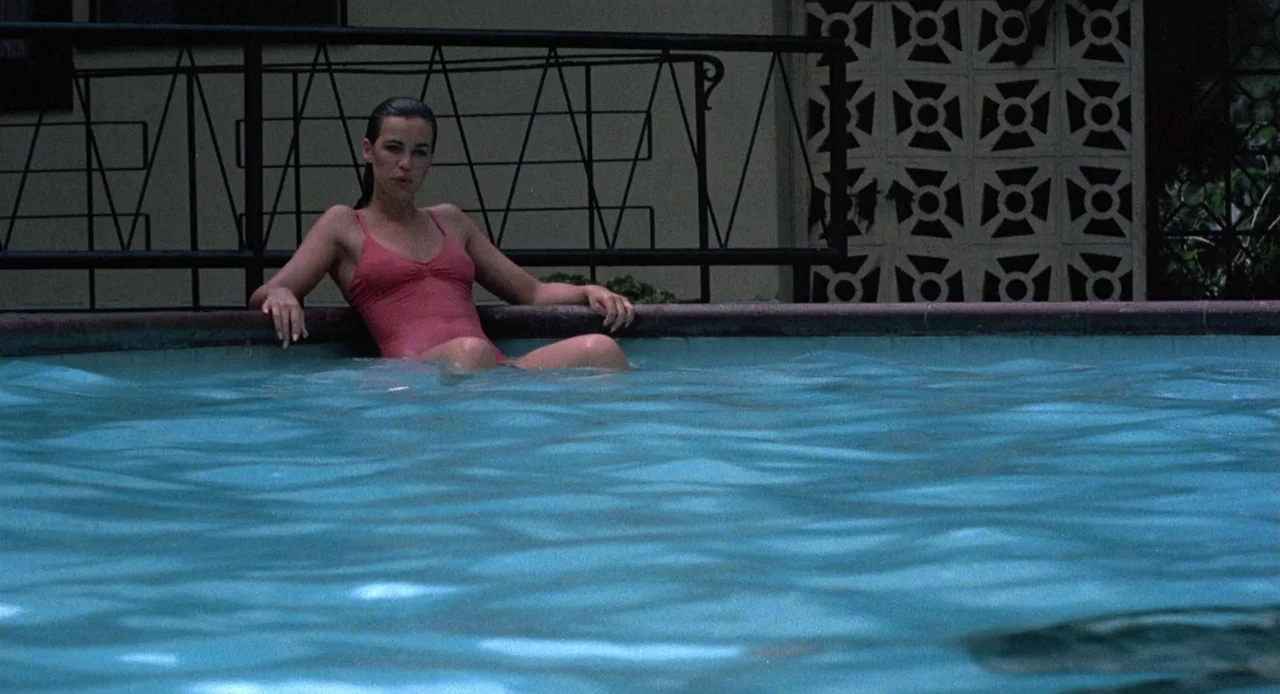 Breathless (1983) movie still: medium — A woman rests on the edge of a turquoise swimming pool in front of a house. The background features a black…