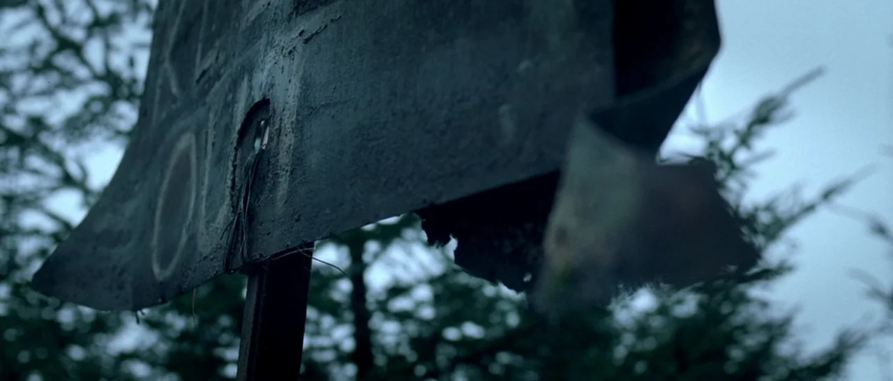 Isolation (2005) movie still: closeup — A weathered, dark metal sign with ragged edges stands against a background of trees. The sign is attached to…
