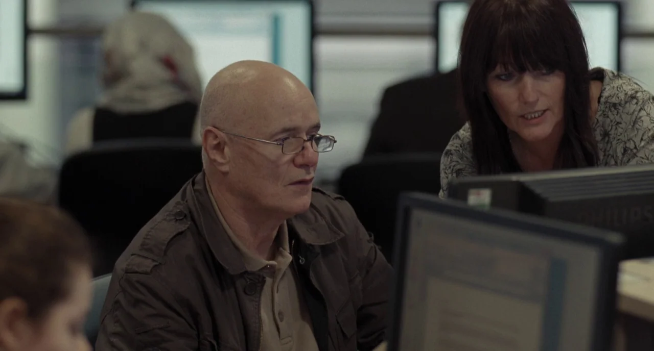 I, Daniel Blake (2016) movie still: medium — An open-plan workplace featuring multiple computer terminals and people working at their desks. A man…