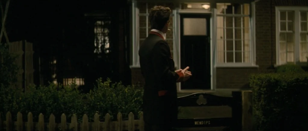 Nowhere Boy movie still: fullbody — A dark residential street at night features a small house behind a wooden fence. A man wearing a dark jacket…