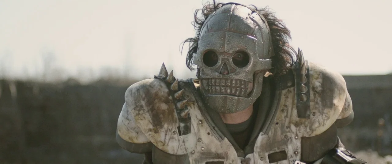 Turbo Kid (2015) movie still: medium — A desolate, overcast outdoor location features a man wearing a metallic skull mask. The person stands in a…