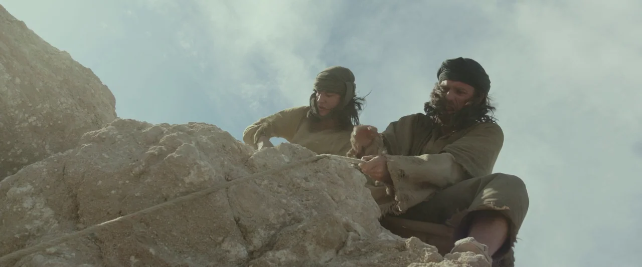 Last Days in the Desert (2015) movie still: medium — Two men in rough ancient tunics struggle with a rope on a rocky cliff. They lean precariously…