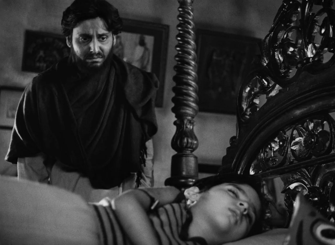 Apur Sansar (1959) movie still: medium — A dimly lit room contains an ornate carved wooden bed with a vertical post. A man stands beside the bed…