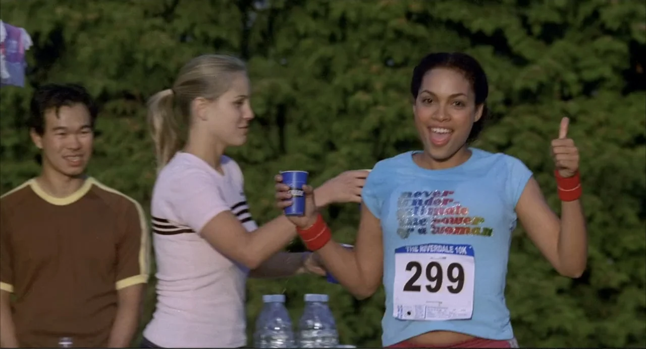 Josie and the Pussycats (2001) movie still: medium — An outdoor race finish line features a young woman smiling and holding a thumb up. Beside her,…