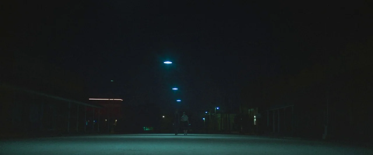 The Vast of Night (2019) movie still: wide — A dark, empty town road is illuminated by a line of overhead streetlights at night. Two figures walk…