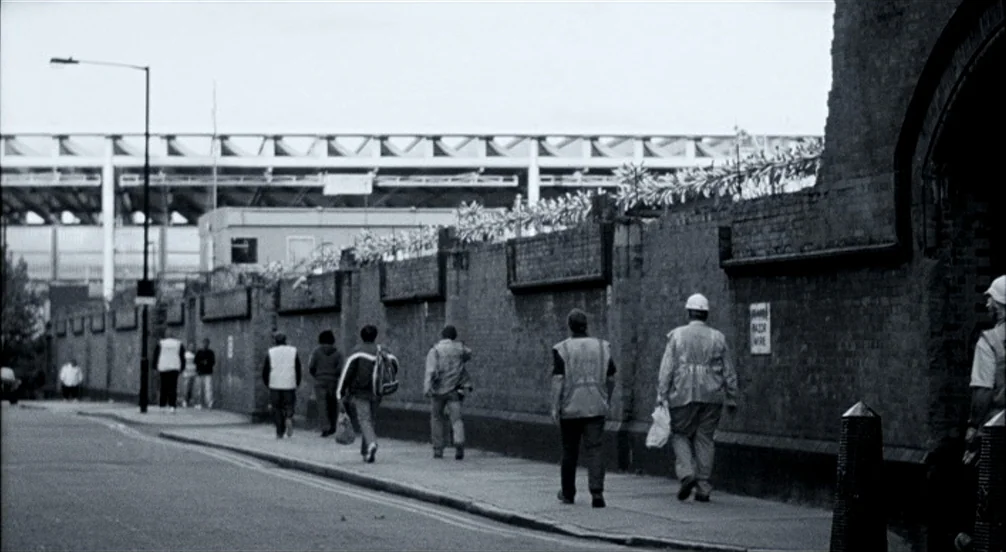 Somers Town (2008) movie still: wide — A long brick wall topped with razor wire runs alongside a paved urban street. A small group of people in work…