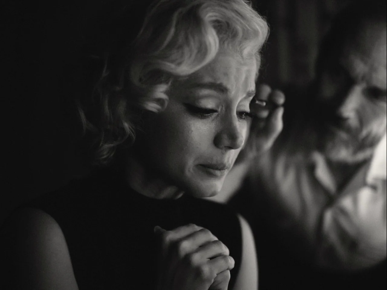 Blonde (2022) movie still: closeup — A dimly lit room with an obscured figure assisting a woman with makeup. The woman has platinum blonde hair and…