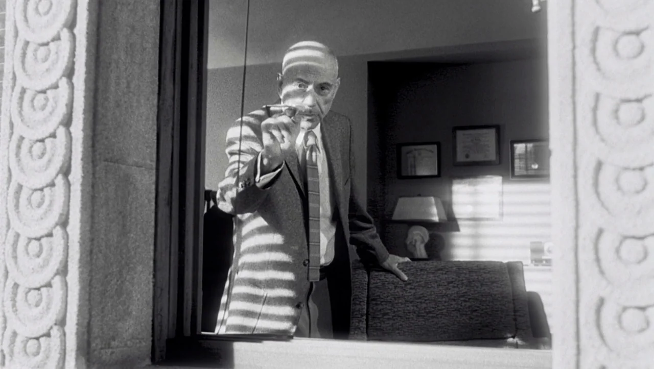 Eros (2004) movie still: medium — A man stands inside a study, viewed through a window frame by the camera. He points a lit cigar directly at the…