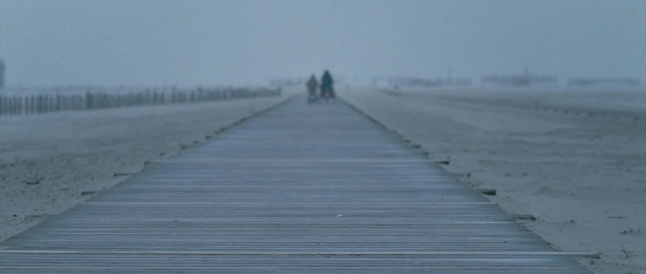 Womb (2010) movie still: wide — A long, straight wooden boardwalk stretches across a flat, sandy, and foggy expanse. Two distant figures walk away…