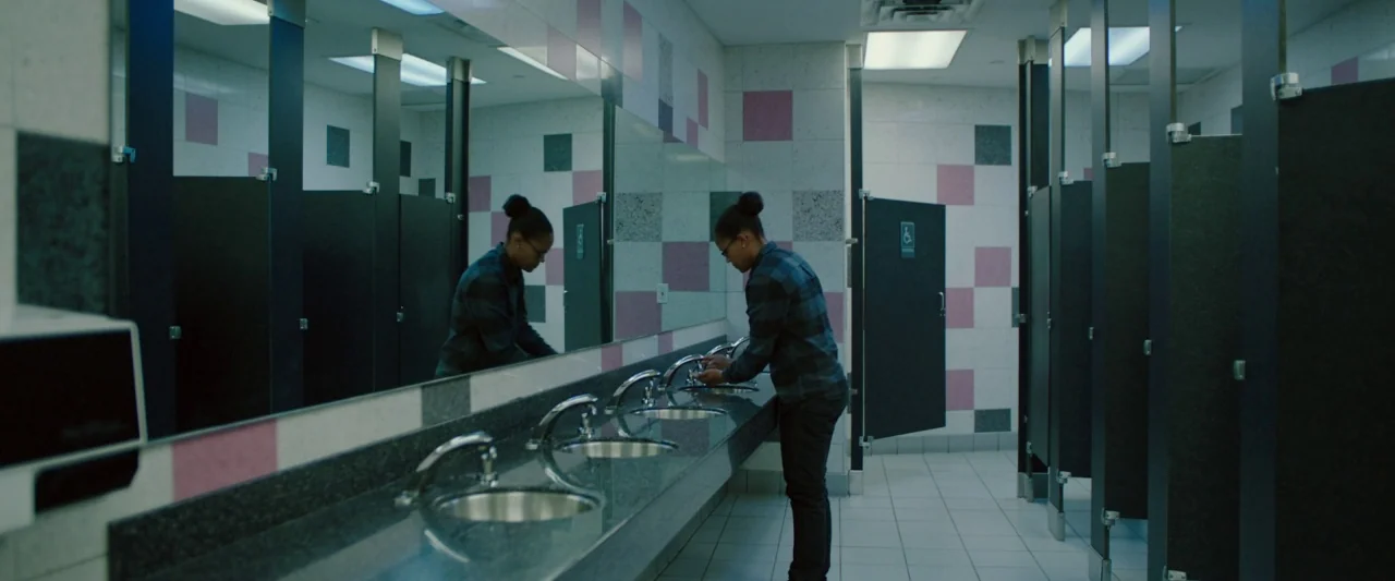 Swallow (2020) movie still: medium — A public restroom interior features tiled walls with alternating pink and grey square accents. A woman stands…
