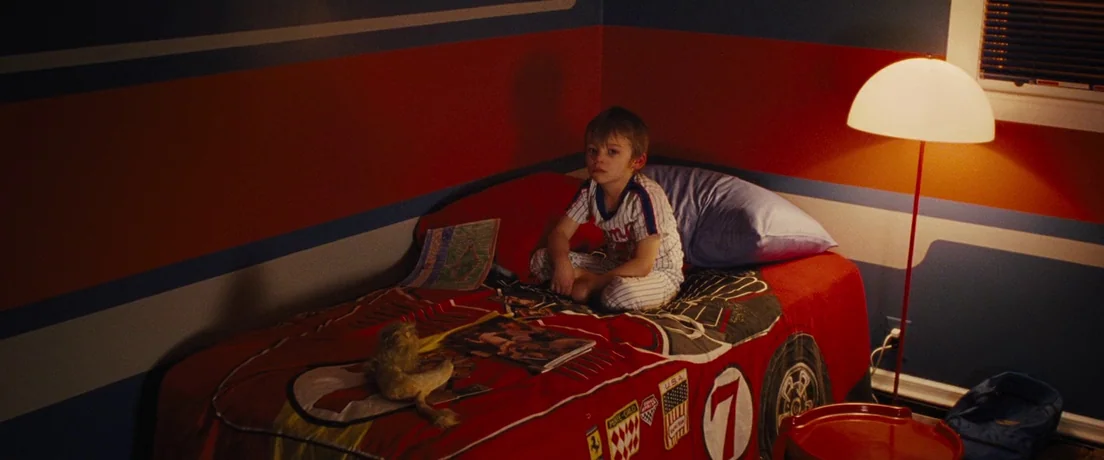 American Hustle (2013) movie still: medium — A dimly lit room features walls with horizontal blue and red stripes. A young boy sits on a…
