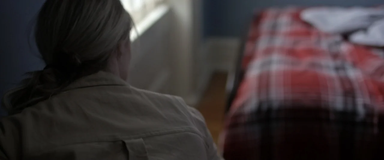 Motherly (2022) movie still: medium — A bedroom contains a bed with a red plaid blanket and a nearby window. A woman in a tan shirt sits on the bed…