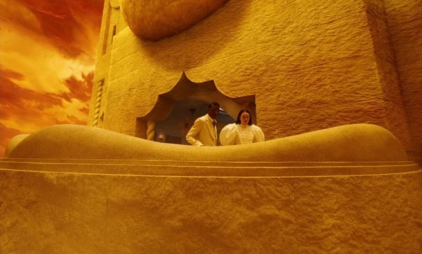 Poor Things (2023) movie still: wide — A stone balcony with carved architectural details overlooks an orange-tinted horizon. A man and a woman stand…