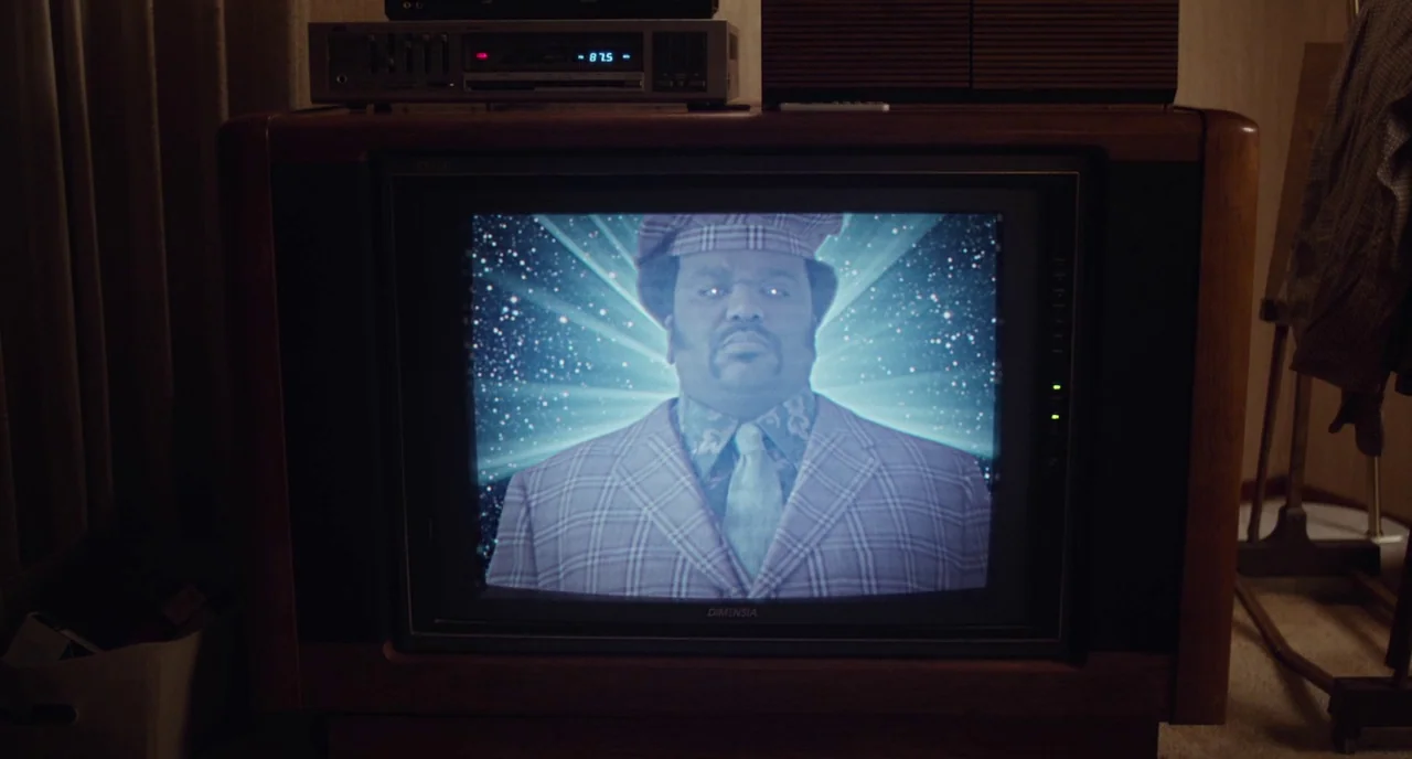 An Evening With Beverly Luff Linn (2018) movie still: closeup — A dark living room contains a large CRT television displaying a man in a blazer. The…