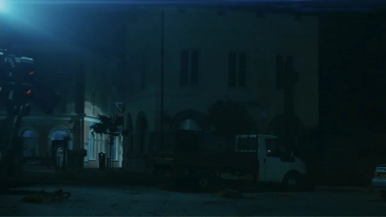Robot Overlords (2015) movie still: wide — An empty nighttime town square features a white truck parked in front of buildings. A lone streetlamp…