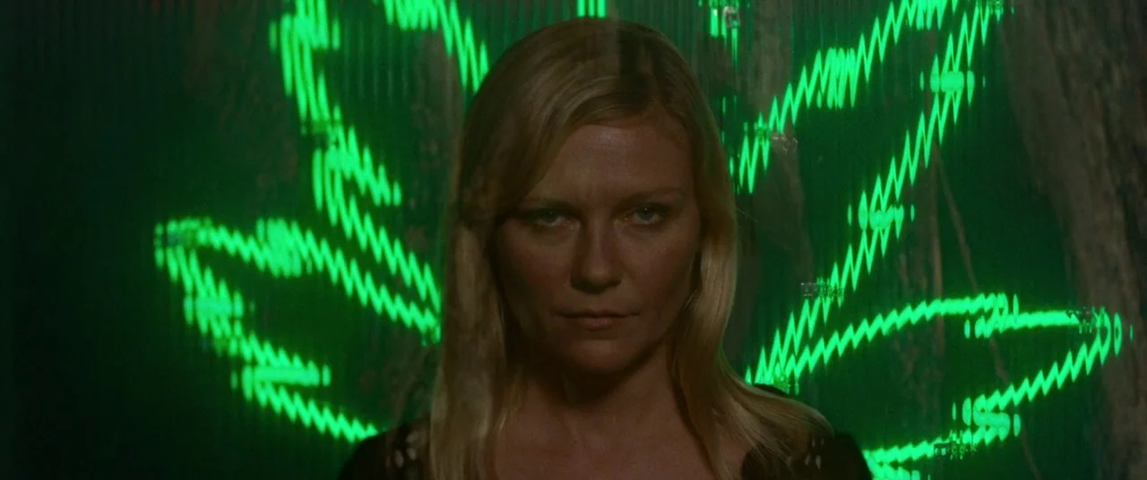Woodshock (2017) movie still: closeup — A woman stands in a dark space against a background of flickering green neon. She stares directly ahead…