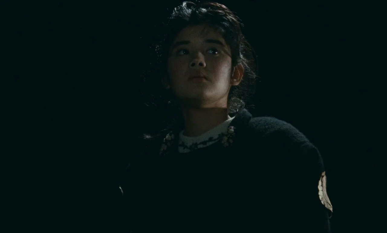 Chizuko’s Younger Sister (1991) movie still: medium — A person stands in total darkness with no visible surroundings or background elements. The…