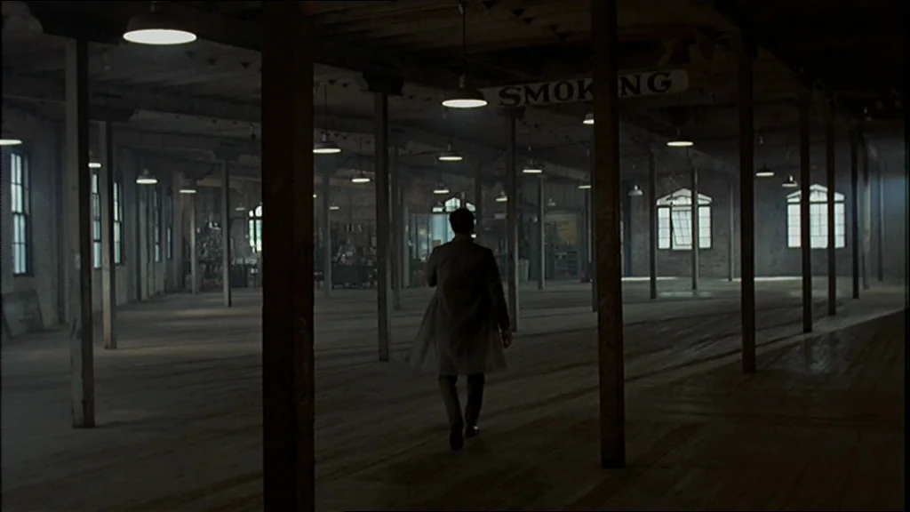 The Conversation (1974) movie still: wide — A vast, empty industrial floor features rows of wooden support pillars and hanging pendant lamps. A man…