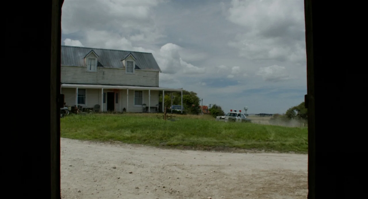 X (2022) movie still: establishing — A light-colored two-story farmhouse sits on a grassy hill under a cloudy sky. A police cruiser is parked on the…