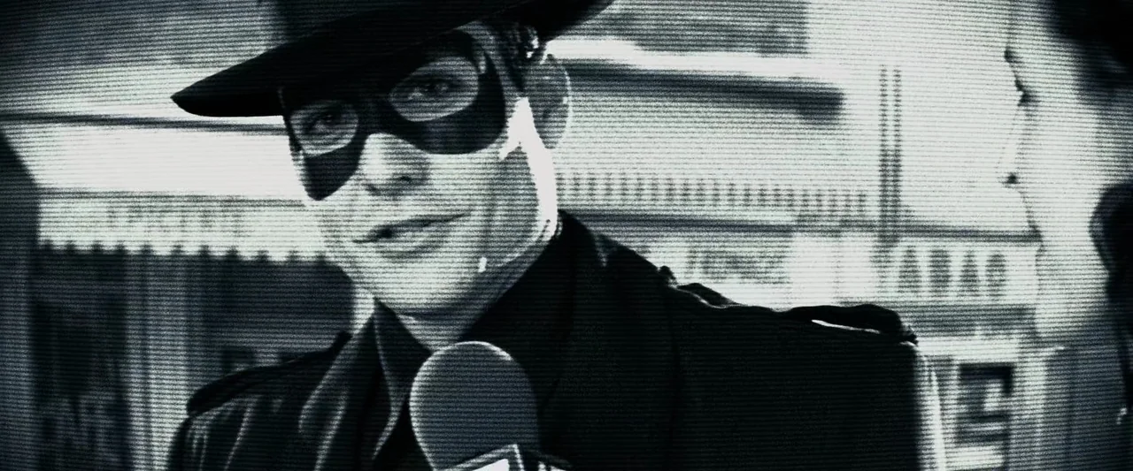 The Spirit (2008) movie still: closeup — A man wearing a fedora and mask speaks into a handheld microphone outside. The grainy footage displays…