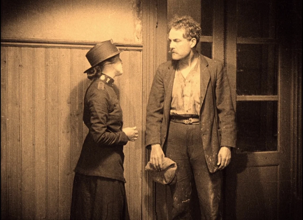 The Phantom Carriage (1921) movie still: medium — A dark hallway with vertical wood paneling and a paneled glass door. A woman in uniform and a…
