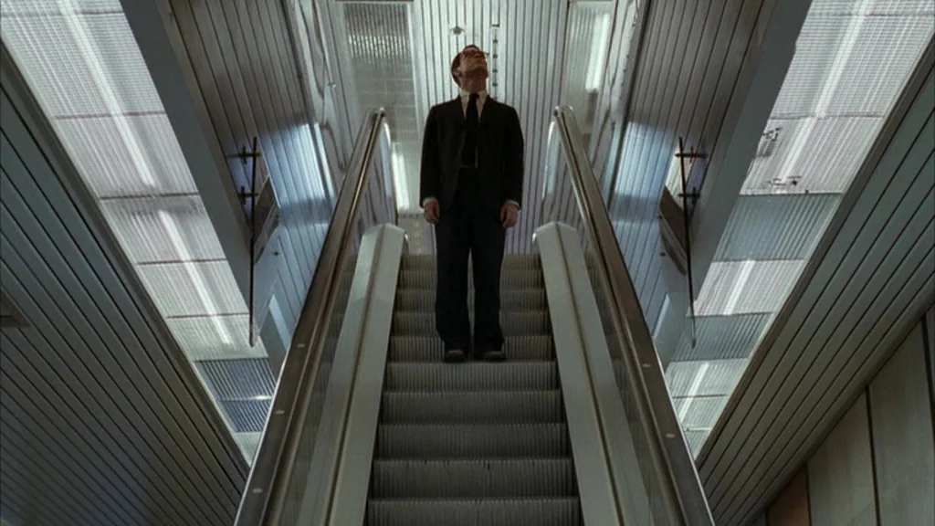 Everything Is Illuminated (2005) movie still: fullbody — An escalator moves upward inside a modern commercial building with ribbed metal walls. A…