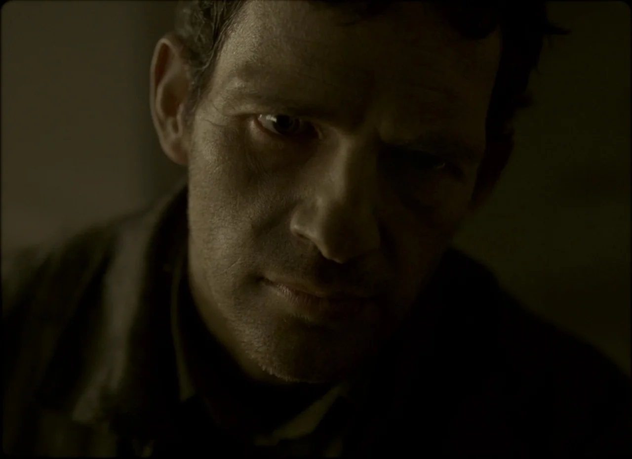 Son of Saul (2015) movie still: closeup — A man stands in a dimly lit, nondescript room with indistinct walls. The man stares directly into the…