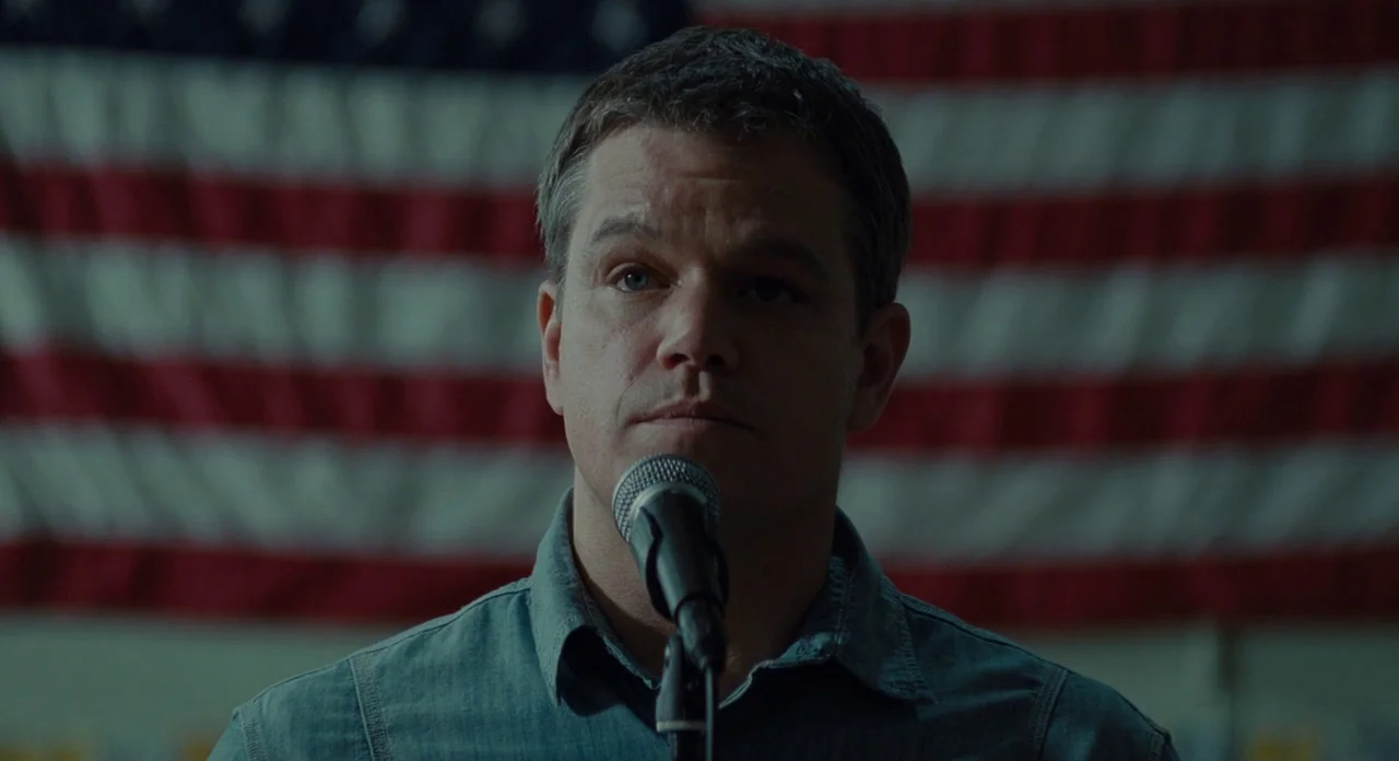 Promised Land (2012) movie still: closeup — A man stands before a large American flag hung against a wall. He speaks into a black microphone…