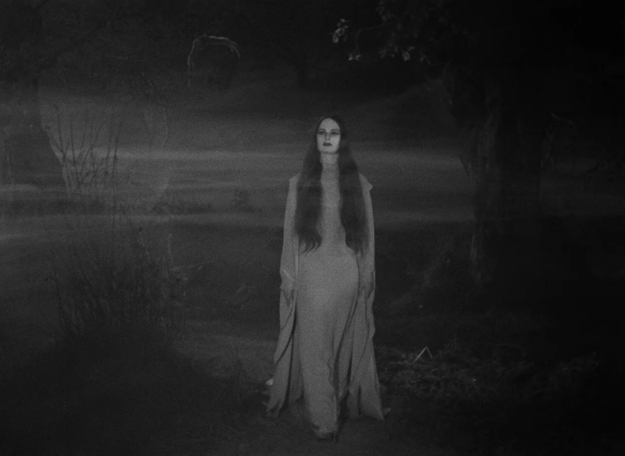 Mark of the Vampire (1935) movie still: fullbody — A woman in a light-colored flowing dress walks through a dark, misty woodland area. The…