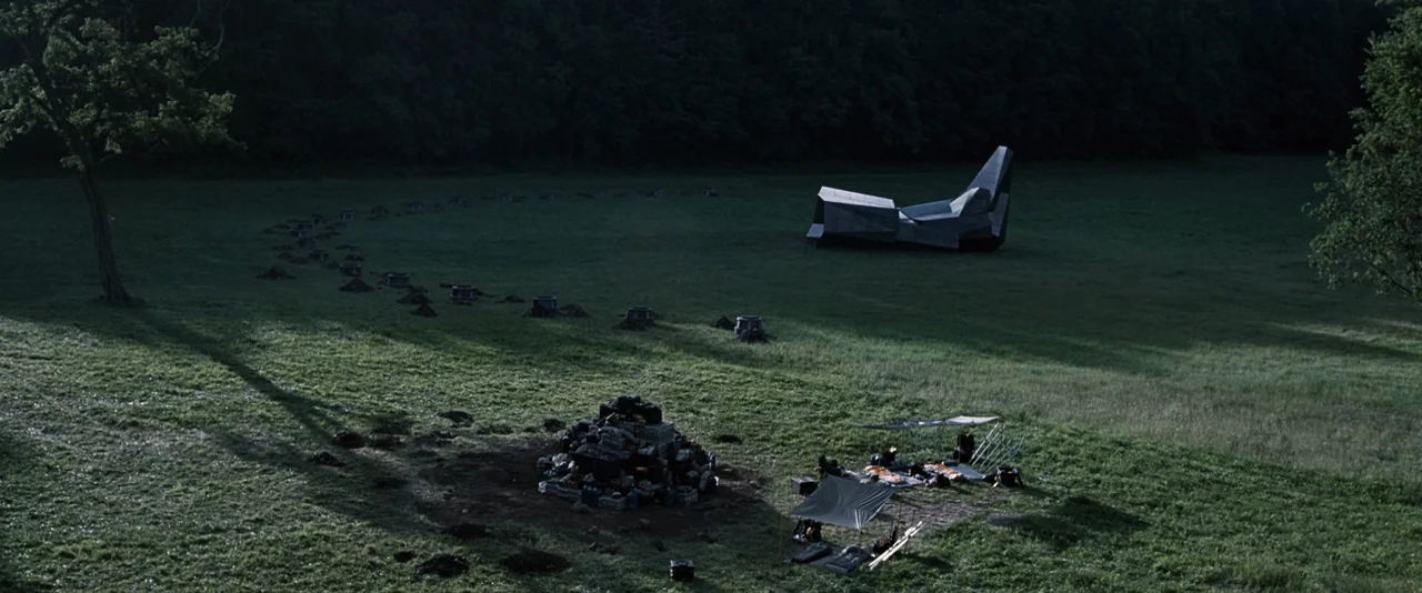 The Hunger Games (2012) movie still: wide — A vast green landscape contains a series of small, dark metallic structures in an arc. A pile of black…
