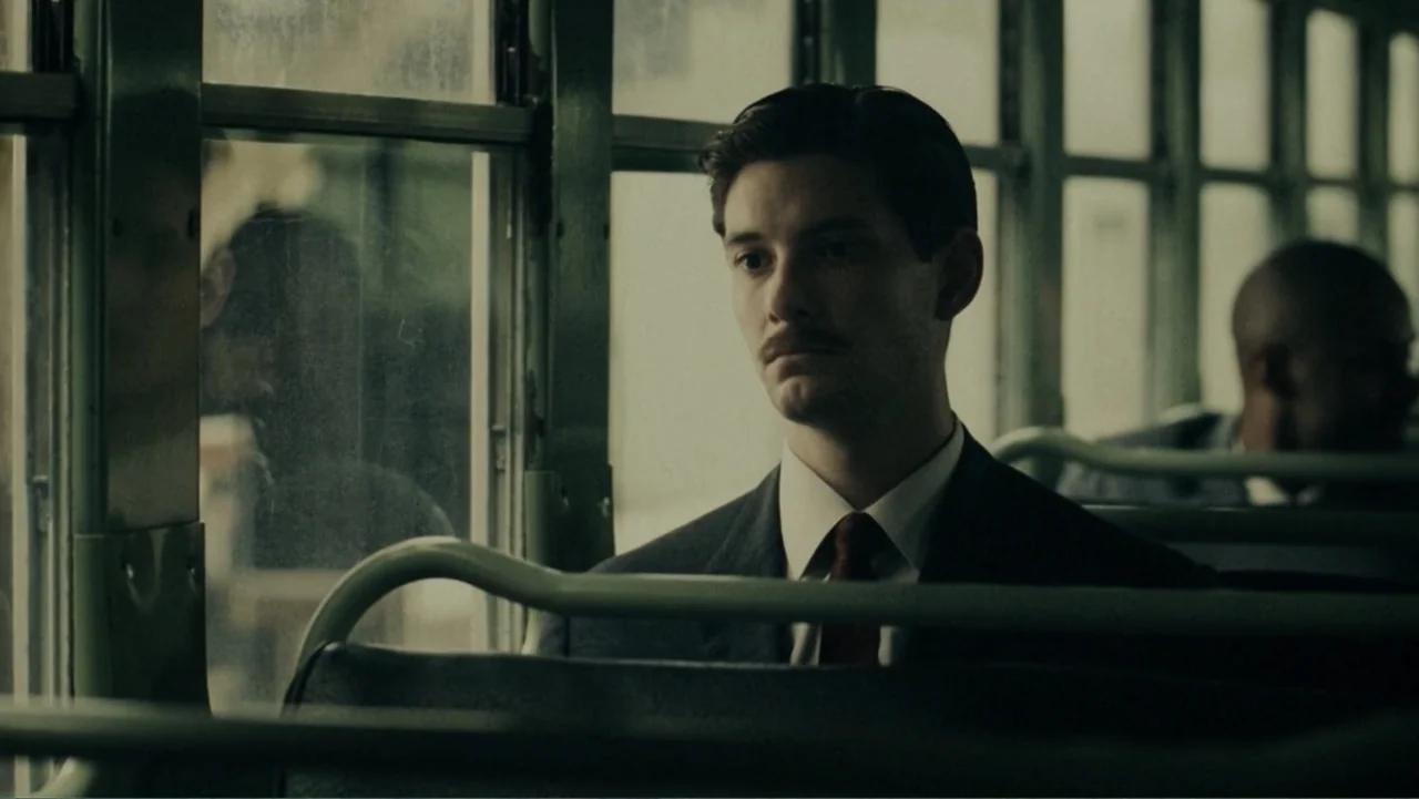The Words (2012) movie still: medium — The interior of a 1950s public bus with metal frame seats and large windows. A man in a suit sits by the…