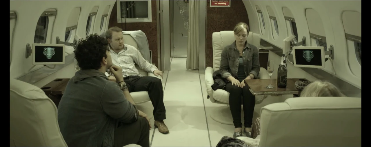 Turbulence (2025) movie still: wide — The interior of a private jet cabin features white leather seats and wood tables. Three individuals sit in…
