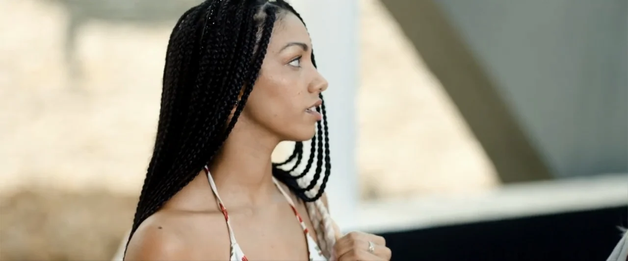 Anaconda (2024) movie still: closeup — An outdoor area with a white structure and blurred sandy background. A young woman with braided hair looks…
