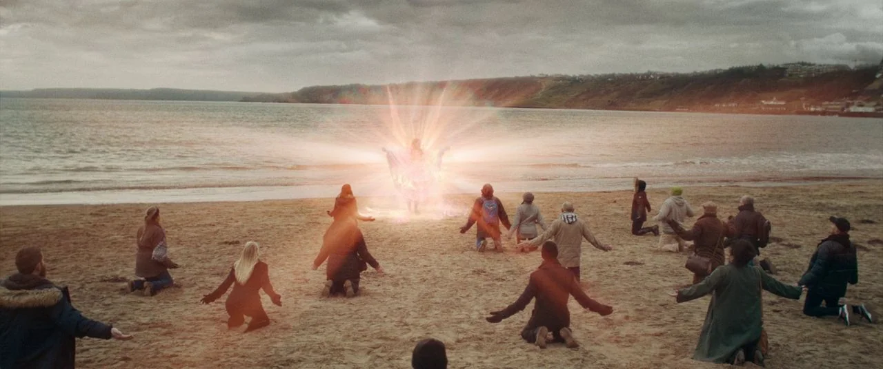 Saint Maud (2019) movie still: wide — A crowd of people kneels on a wide sand beach under an overcast sky. A glowing, radiating humanoid figure…