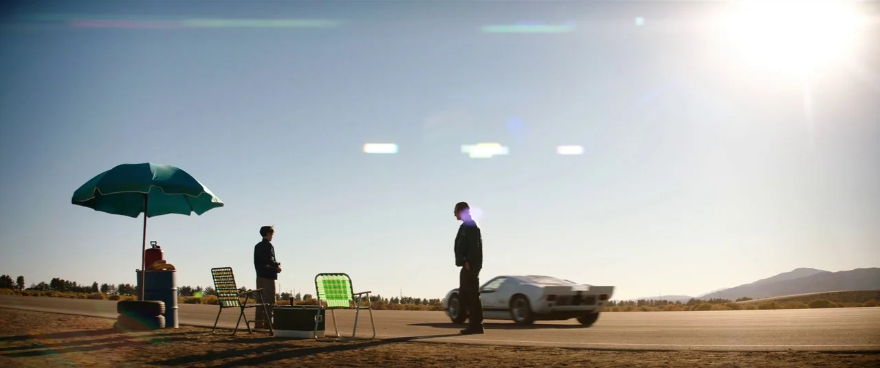 Ford V Ferrari (2019) movie still: wide — A vast, empty asphalt runway sits under a bright, clear sunlit sky. Two men stand apart near a small setup…