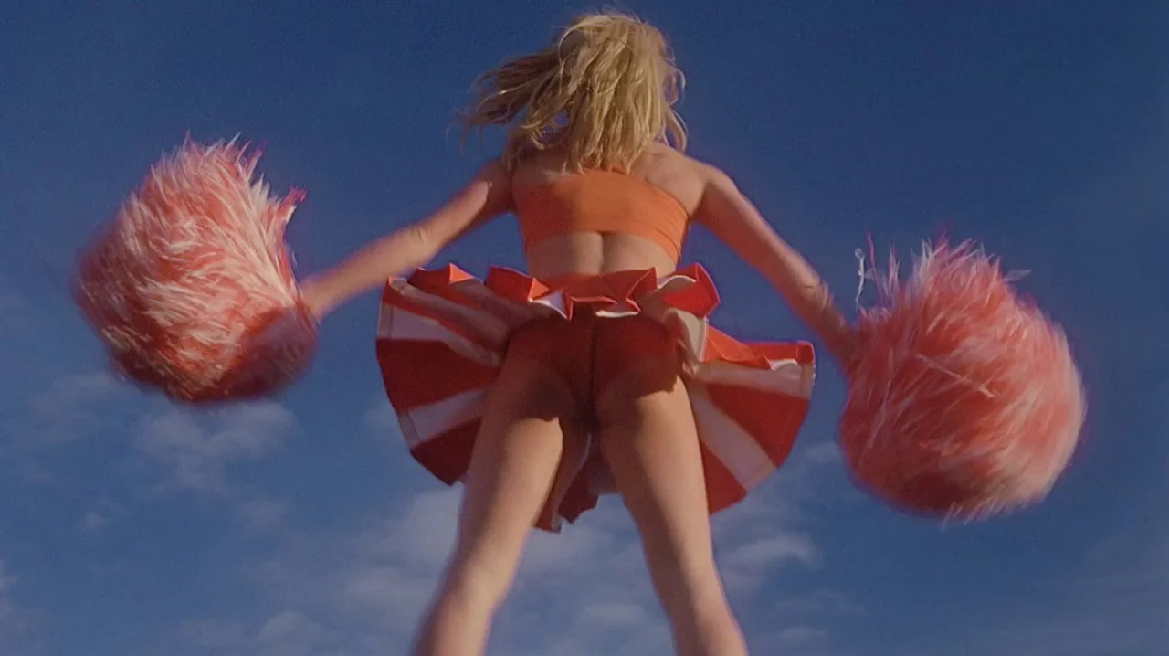 But I’m A Cheerleader (1999) movie still: fullbody — A cheerleader performs against a clear blue sky. She holds orange pom-poms while wearing a…