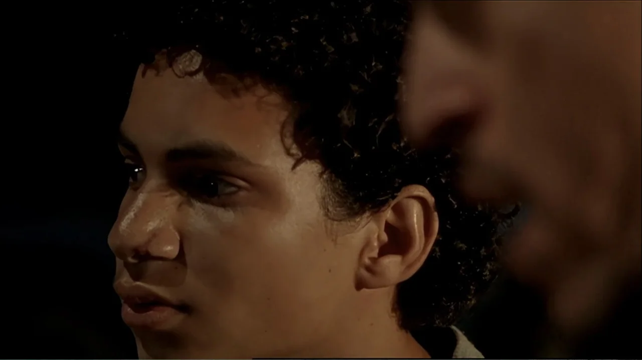 In the Middle of the World (1990) movie still: closeup — A dark, nondescript interior space frames a young man in profile from close proximity. The…