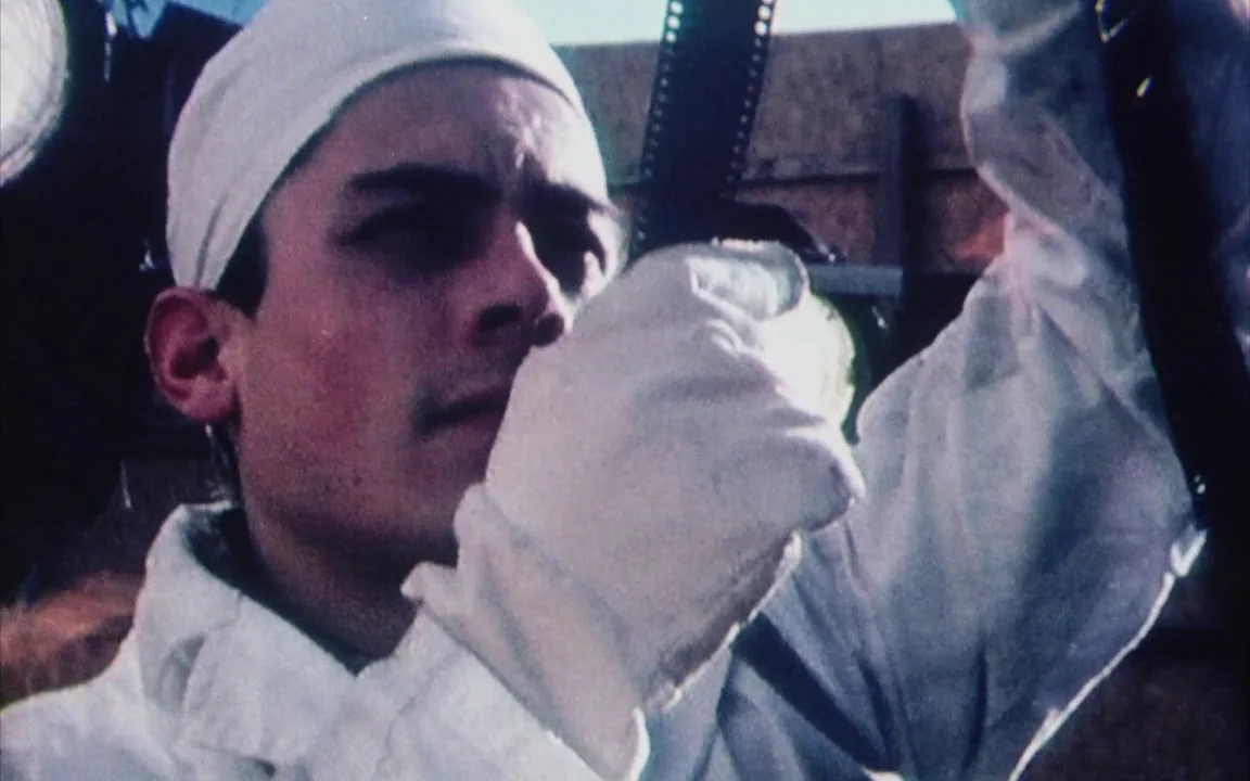 The Last of England (1987) movie still: closeup — A man in a white lab coat examines a piece of film stock. He wears protective gear in a bright…