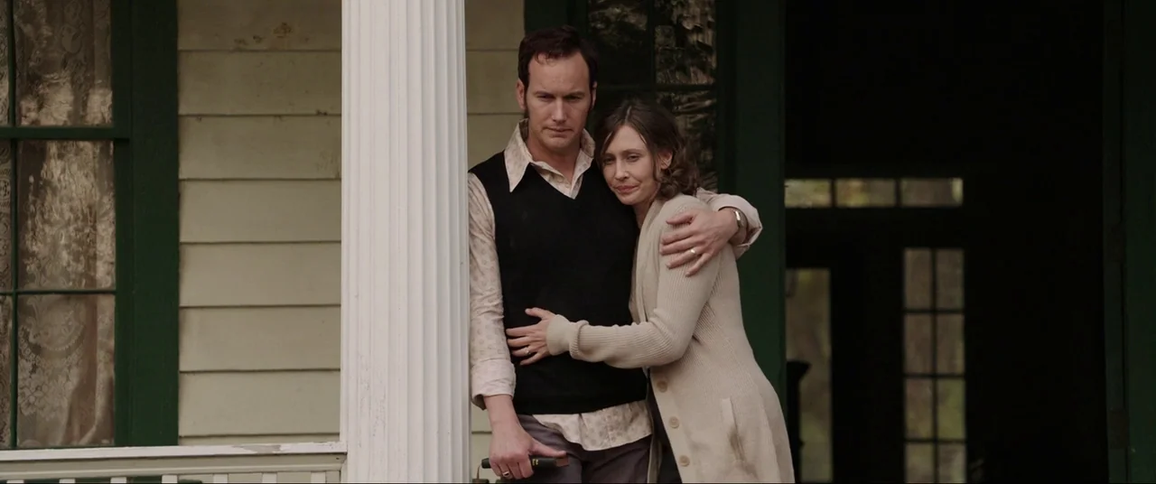 The Conjuring (2013) movie still: medium — A wooden porch with a white column stands in front of a house exterior. A man and a woman hold each other…