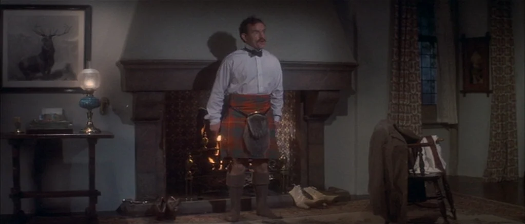 The Private Life of Sherlock Holmes movie still: fullbody — A dimly lit room contains a stone fireplace with a fire burning inside. A man wearing a…