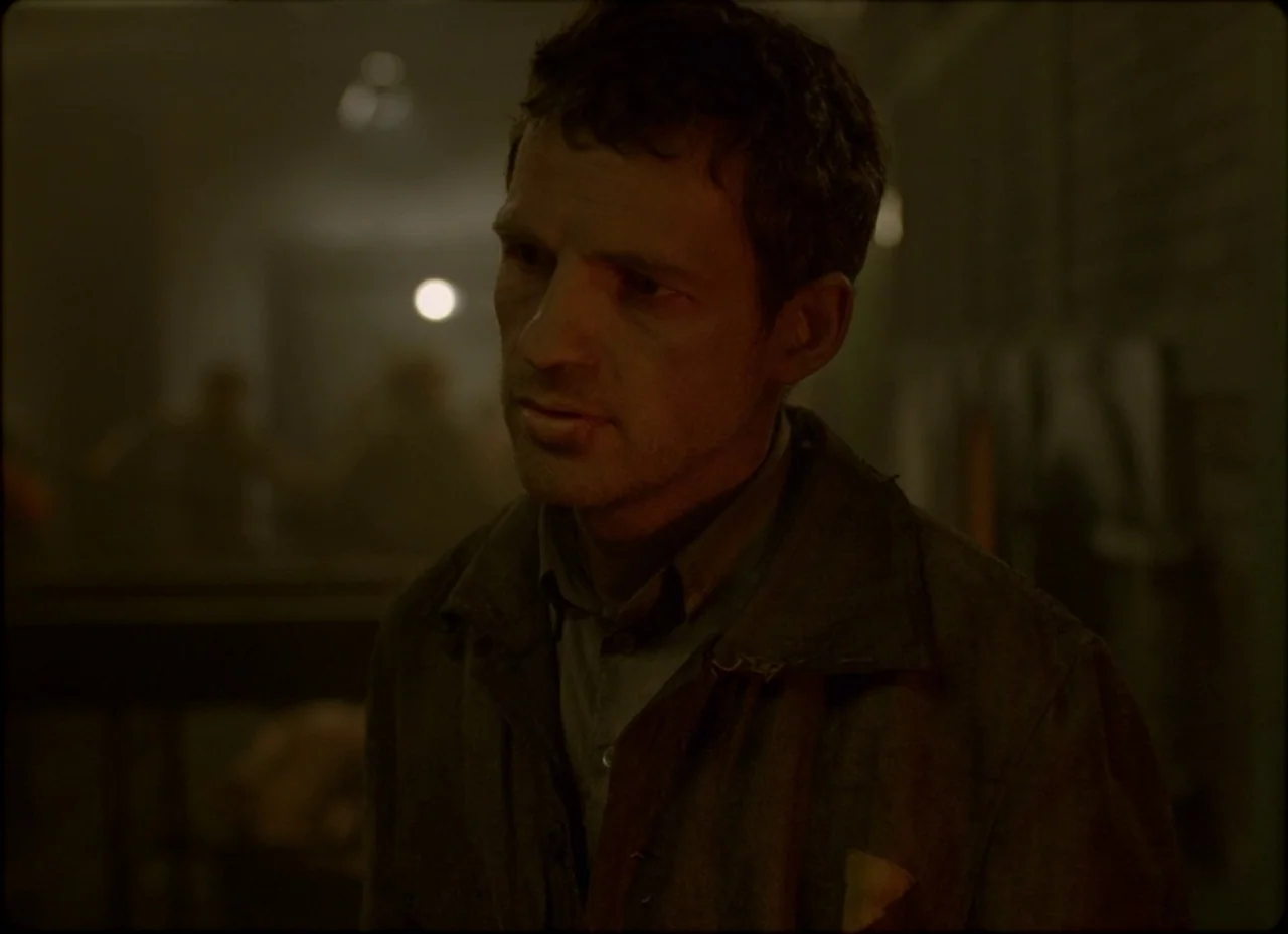 Son of Saul (2015) movie still: medium — A dimly lit, dusty workshop space contains indistinct silhouettes in the blurred background. A man stands…