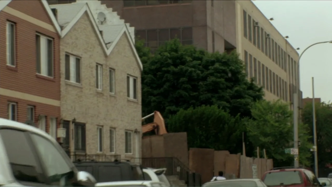 Little White Lie (2008) movie still: wide — A quiet urban street shows residential townhouses beside a large apartment building. Vehicles line the…