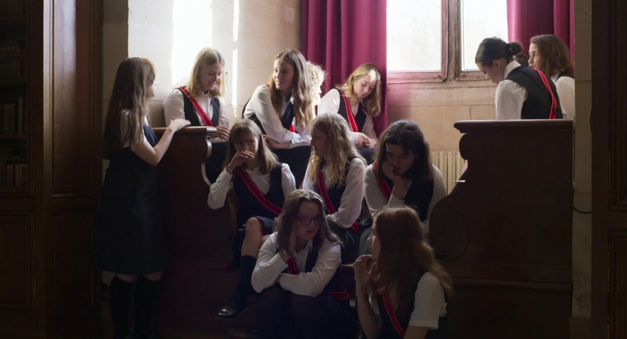 Zombi Child (2019) movie still: wide — A group of teenage girls in uniform gather on wooden benches inside a library. They are seated in casual…