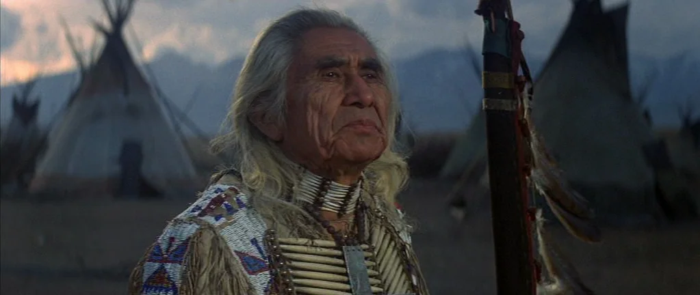 Little Big Man (1970) movie still: medium — An indigenous man stands in an outdoor encampment with several conical tents visible behind. He looks…