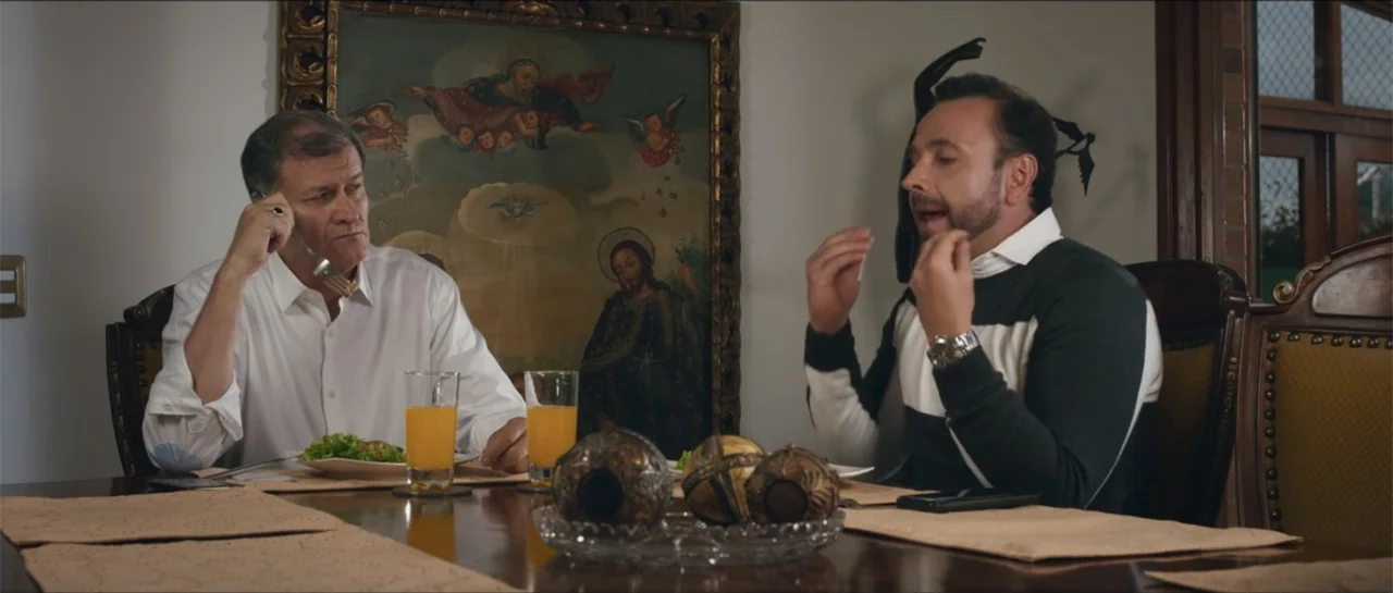 Loving is Losing (2019) movie still: medium — A formal dining room features a large religious painting on the wall behind them. Two men are seated…