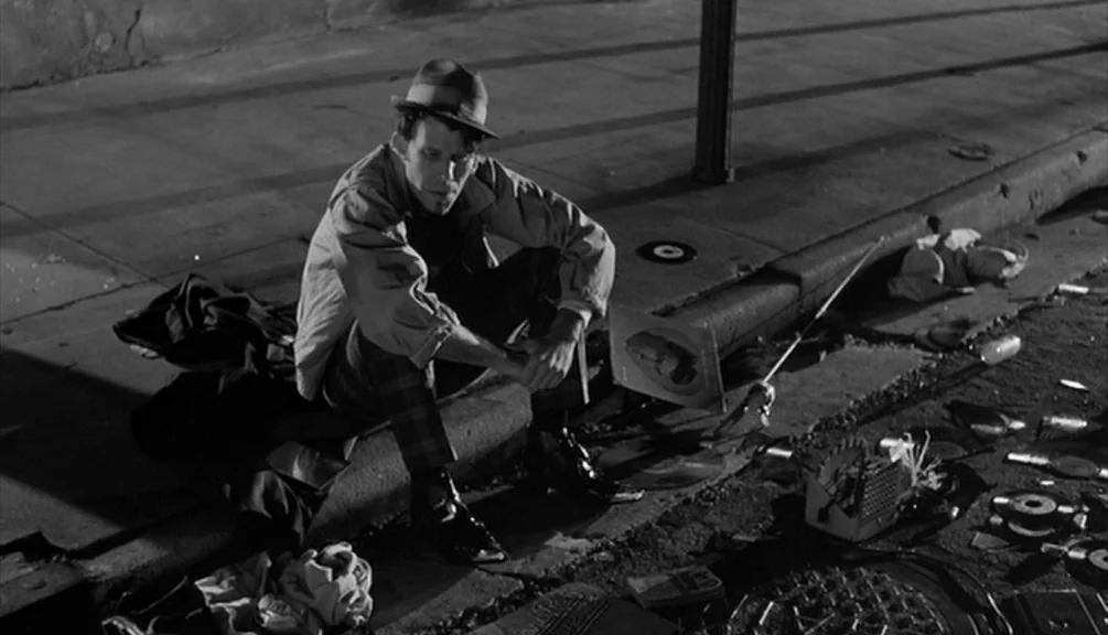 Down By Law (1986) movie still: medium — A man sits on a concrete curb at night surrounded by scattered vinyl records. The pavement is lit by harsh…