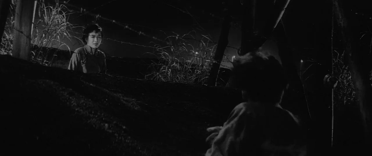 The Human Condition I: No Greater Love (1959) movie still: medium — An embankment at night is divided by a line of barbed wire fencing. A woman…