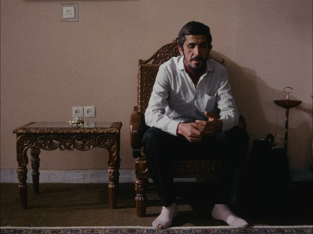 Close-Up (1990) movie still: medium — A man sits on a carved wooden chair against a plain wall. A small ornamental table and a floor ashtray flank…