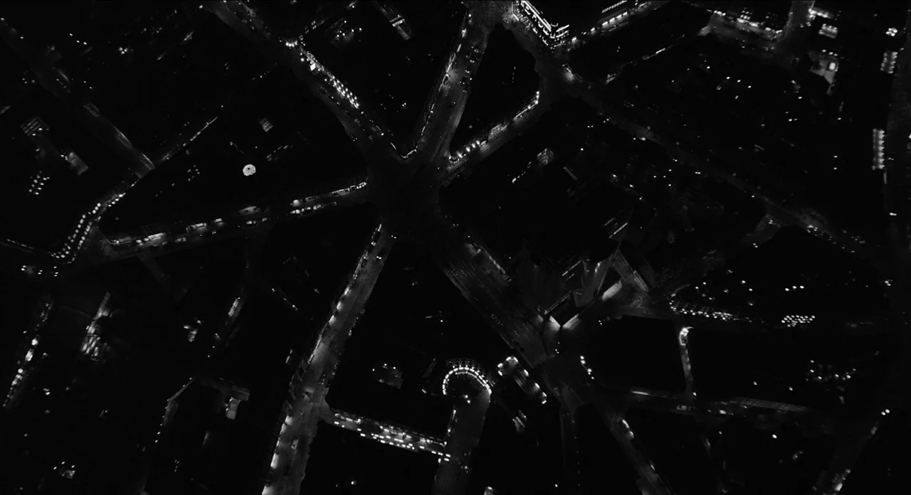 One More Time With Feeling movie still: establishing — An overhead aerial view depicts a dark urban grid with intersecting roads at night. Small…